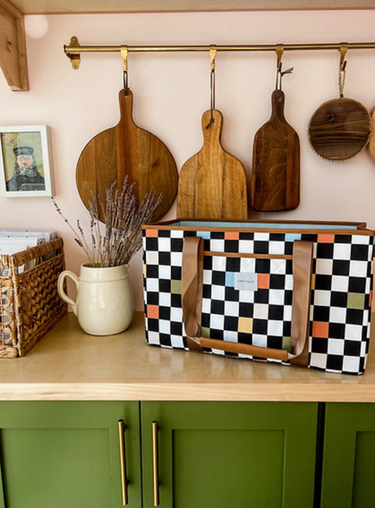 A patterned fabric tote bag with brown straps sits on a wooden kitchen counter. The bag's black and white checkerboard pattern features squares of light blue, tan, and orange. In the background, there are various wooden cutting boards hanging on a wall.